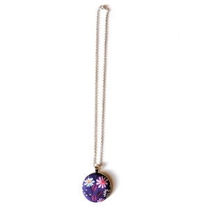 The circle shaped pendant hangs from a silver colored chain. The pendant is purple with pink, black, and white flowers and leaves.