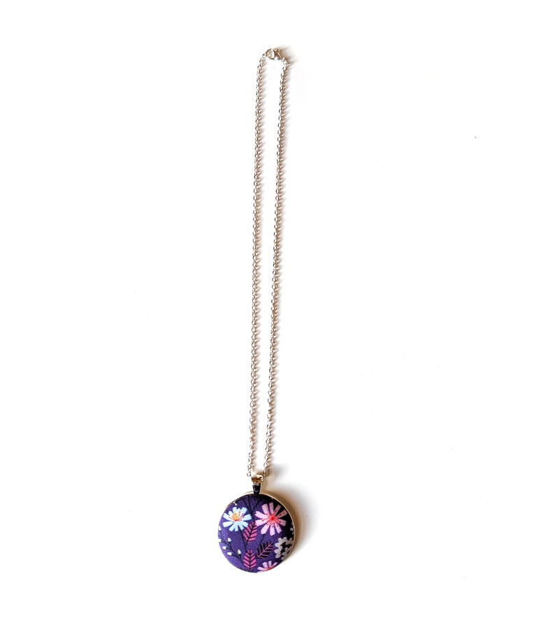 The circle shaped pendant hangs from a silver colored chain. The pendant is purple with pink, black, and white flowers and leaves.