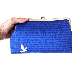 The model's hand is holding out the clutch. The back of the clutch is shown. It is blue with dark blue text. There is a single white bird near the bottom.