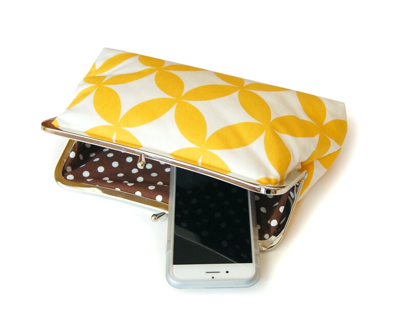 The yellow and white clutch is shown on its side, slightly opened. Inside there is a brown and white polka dot print. There is a phone resting inside the bag.