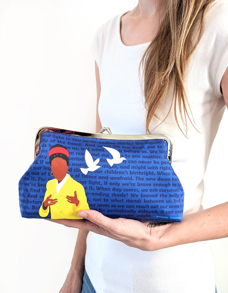 The model is holding the Amanda Gorman clutch purse in front of her.