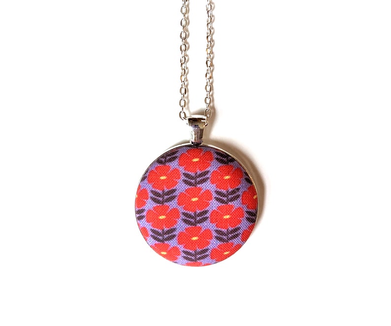 The fabric pendant necklace is hanging from a silver colored chain. The pendant has a light purple background with a pattern of red flowers.