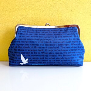 The back of the clutch is shown. It is blue with dark blue text. There is a single white bird near the bottom. The clutch is on a white table with a yellow wall behind it.