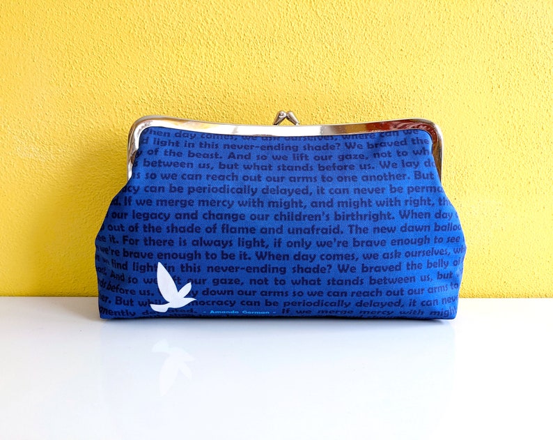 The back of the clutch is shown. It is blue with dark blue text. There is a single white bird near the bottom. The clutch is on a white table with a yellow wall behind it.