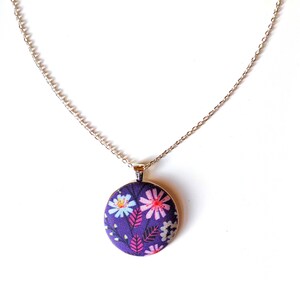 The circle shaped pendant hangs from a silver colored chain. The pendant is purple with pink, black, and white flowers and leaves.