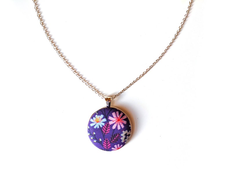 The circle shaped pendant hangs from a silver colored chain. The pendant is purple with pink, black, and white flowers and leaves.