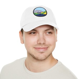 May include: A white baseball cap with a circular patch featuring a landscape scene with mountains, a river, and green fields.