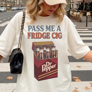 May include: Cream-colored t-shirt with the text "PASS ME A FRIDGE CIG" in blue and orange. The shirt features a graphic of a Dr. Pepper box with bottles inside. A beige baseball cap with a small blue logo is worn.