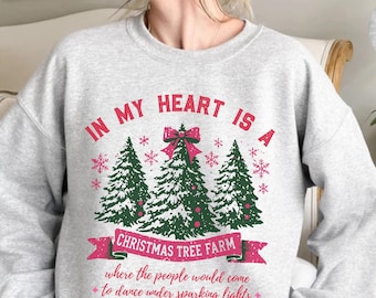 Op de afbeelding: Een grijs sweatshirt met de tekst "IN MY HEART IS A CHRISTMAS TREE FARM where the people would come to dance under sparking lights" gedrukt op de voorkant. De tekst is in rood en roze met een groene en witte illustratie van drie kerstbomen.