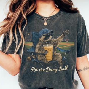 Raccoon wearing sunglasses sitting in a folding chair holding iced coffee at a baseball field with humorous sports quote on casual t-shirt