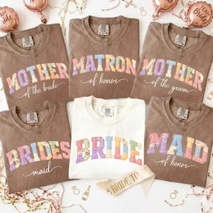 May include: A collection of brown and white t-shirts with text for a bridal party. The shirts say "Mother of the Bride", "Matron of Honor", "Mother of the Groom", "Bridesmaid", "Bride", and "Maid of Honor". Balloons and a sash are also visible.