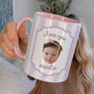 May include: A white ceramic mug with a pink handle and interior. The mug features a photo of a baby with the words "i love you auntie" in purple script. The mug has a striped pattern in white and lavender.