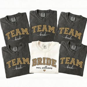 May include: A collection of gray and white t-shirts with leopard print lettering. The gray shirts say "TEAM bride" and the white shirt says "BRIDE mrs. williams" with a heart graphic. The shirts are likely for a bridal party.