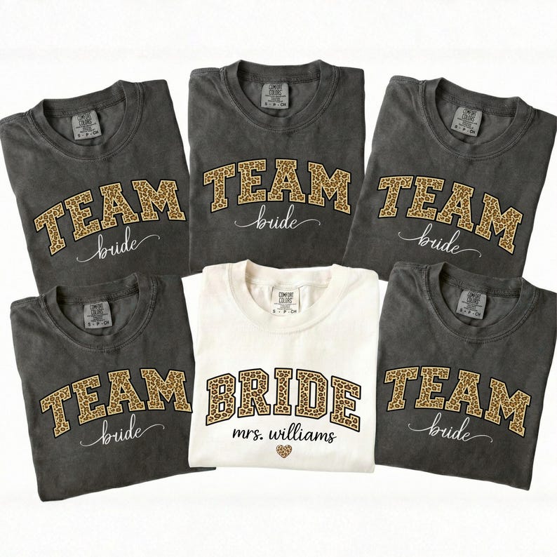 May include: A collection of gray and white t-shirts with leopard print lettering. The gray shirts say "TEAM bride" and the white shirt says "BRIDE mrs. williams" with a heart graphic. The shirts are likely for a bridal party.