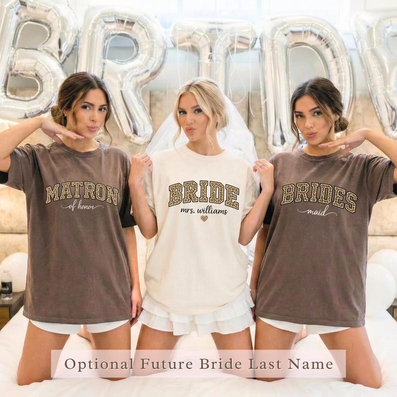 May include: Three women wearing matching t-shirts for a bridal party. The shirts are brown and cream-colored, with text that reads "Matron of Honor", "Bride", and "Bridesmaid". The word "Bride" includes the text "Mrs. Williams".