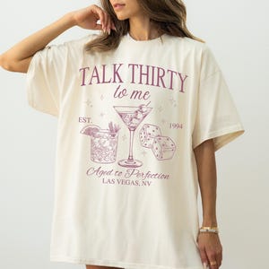 Puede incluir: Una camiseta blanca con un dise&ntilde;o gr&aacute;fico rosa que dice "Talk Thirty to Me" con una copa de martini y dados. El gr&aacute;fico tambi&eacute;n dice "Est. 1994" y "Aged to Perfection Las Vegas, NV".