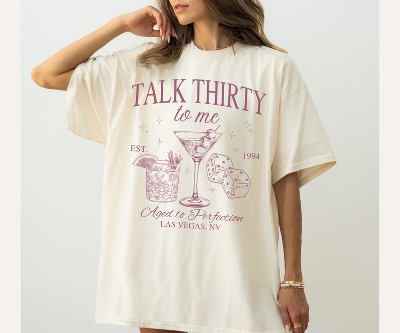 Puede incluir: Una camiseta blanca con un dise&ntilde;o gr&aacute;fico rosa que dice "Talk Thirty to Me" con una copa de martini y dados. El gr&aacute;fico tambi&eacute;n dice "Est. 1994" y "Aged to Perfection Las Vegas, NV".