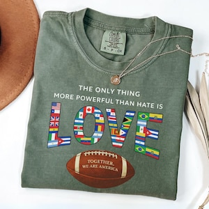 May include: Olive green t-shirt with the text "THE ONLY THING MORE POWERFUL THAN HATE IS LOVE". The word "LOVE" is filled with various country flags. A brown football with the text "TOGETHER, WE ARE AMERICA" is below the word "LOVE".