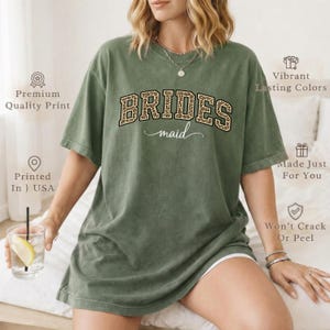 May include: An olive green t-shirt with the words "BRIDES maid" in a leopard print design. The shirt has short sleeves and a relaxed fit. The text "Printed In USA" is also visible.