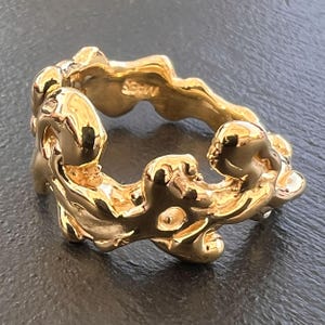 Può includere: Un anello in oro con un design unico, organico e testurizzato. L'anello è realizzato in un pezzo spesso e solido di metallo e ha una superficie ruvida e irregolare.