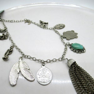 Op de afbeelding: Een zilveren ketting met meerdere bedels, waaronder een veer, een schildpad, een turquoise steen en een religieuze medaille. De ketting heeft ook een kwast aan het einde.