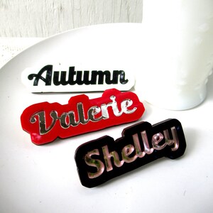 Pode incluir: Três placas de nome personalizadas com acabamento espelhado. Os nomes são "Autumn", "Valerie" e "Shelley". As placas são feitas de acrílico e têm um fundo branco, vermelho e preto.