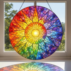 May include: A round stained glass window with a vibrant rainbow design. The artwork features a central yellow sunburst radiating outwards through shades of red, orange, yellow, green, blue, and purple. The window is hanging in front of a window.