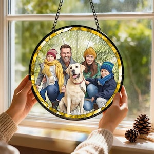 May include: A round, stained-glass window art piece featuring a family portrait with a dog. The artwork is held up by a chain, showcasing a family of four and a yellow Labrador Retriever. The window has a black border and yellow stained glass.