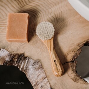 May include: A wooden brush with natural bristles and a wooden handle. The handle has the text "hydréa london" printed on it. The brush is on a wooden surface next to a bar of soap.