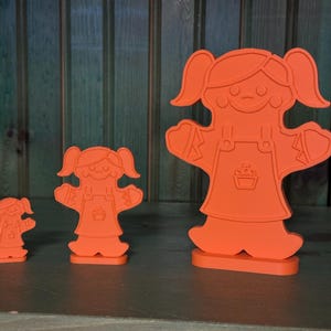 May include: Three orange plastic figurines of people with pigtails and dresses. The figurines are different sizes, with the largest figurine being about 10 inches tall.