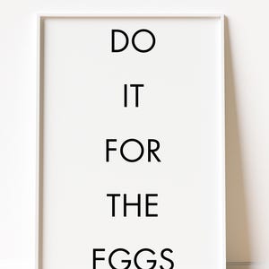 May include: A framed white print with the text "DO IT FOR THE EGGS" in a bold, black font. The artwork is displayed against a white wall and rests on a light-colored wooden floor.