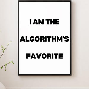 May include: A framed art print with the text "I AM THE ALGORITHM'S FAVORITE" in bold black letters on a white background. The print is in a black frame and is displayed on a white wall.