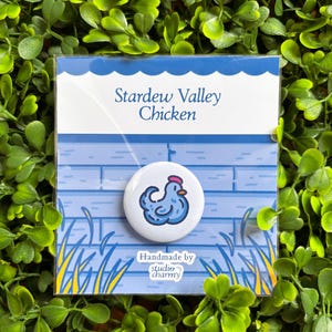 Könnte beinhalten: Ein weißer Ansteckbutton mit einem blauen Cartoon-Huhn und rotem Kamm. Der Button befindet sich auf einer Karte mit dem Text "Stardew Valley Chicken" und "Handmade by studio charmy". Der Hintergrund hat ein Backsteinmauer-Design.