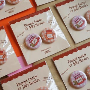 Peut inclure: Plusieurs badges avec l'inscription "Peanut butter & Jelly Besties" sont présentés. Chaque badge représente un dessin animé d'un pot de confiture et d'un pot de beurre de cacahuète. Les badges sont emballés sur des cartes.