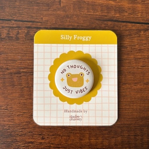 Könnte beinhalten: Ein Ansteckbutton mit einem Frosch-Cartoon-Design und dem Text "NO THOUGHTS JUST VIBES". Der Button befindet sich auf einer gelben Karte mit der Aufschrift "Silly Froggy" und "Handmade by studio charmy".