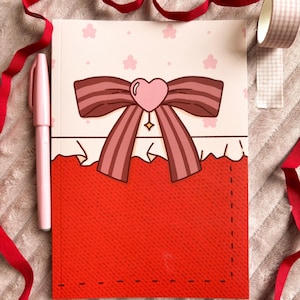 Pode incluir: Um caderno com capa vermelha e um laço decorativo com um coração no centro. Uma caneta rosa e uma fita vermelha são colocadas ao lado do caderno. Um rolo de fita adesiva estampada também é visível.
