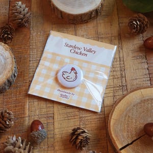 Op de afbeelding: Een button met een cartoon kip illustratie, verpakt op een gele ruitjeskaart. De tekst "Stardew Valley Chicken" staat boven de button. De button is "Handmade by studio charmy". De achtergrond is een houten oppervlak met dennenappels en eikels.