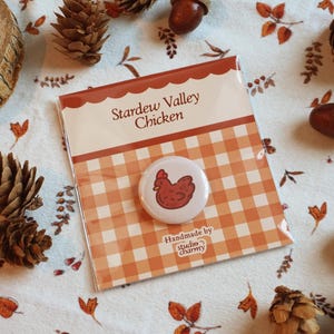 Könnte beinhalten: Ein Ansteckbutton mit einer roten Hühnerillustration, verpackt auf einer Karte mit dem Text "Stardew Valley Chicken". Die Karte hat ein orange-weißes Vichy-Muster. Der Button ist "Handmade by studio charmy".