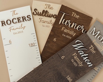 May include: A collection of wooden growth charts with family names and the year established. The charts are in various colors, including white, brown, and gray. Each chart has a measurement scale in inches and the text "The [Family Name] Family Est. [Year]"