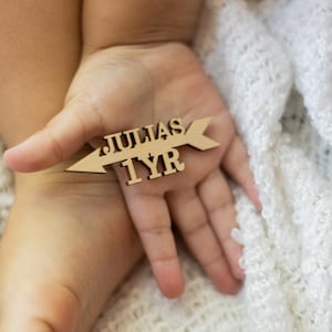 以下が含まれることがあります： 「JULIAS」と「1YR」の文字が刻まれた、矢印型の木製記念品。柔らかい白い毛布を背景に、人の手の中に矢印が収まっています。矢印は明るい色の木材で作られています。