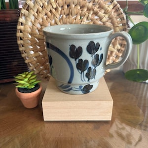 May include: A ceramic mug with a light blue glaze, adorned with dark blue floral patterns and a blue stripe. The mug has a handle and sits on a small wooden block. A small succulent in a terracotta pot is in the foreground.