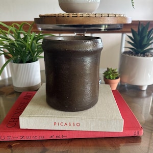 May include: A cylindrical dark brown ceramic vessel with a textured, matte finish and a slightly flared rim. The vessel is positioned on top of a light beige book titled Picasso, which rests on a larger red book titled Chinese Art.