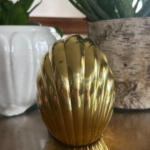 May include: A polished brass decorative object shaped like a seashell with vertical fluted ridges. The surface is smooth and reflective, catching light across its curved, rounded form. The object has a symmetrical design with a pointed top.