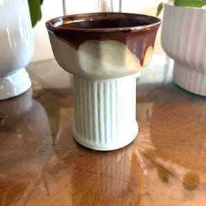May include: A ceramic pedestal bowl with a brown rim and interior, a cream-colored exterior, and a white, ribbed pedestal base. The bowl sits on a wooden surface, with other decorative items in the background.