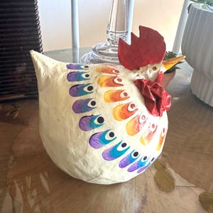May include: A handcrafted, off-white chicken figurine with colorful, stylized feather designs in shades of purple, blue, orange, and yellow. The chicken has a red comb and wattle, and a yellow beak. The figurine is a decorative item.