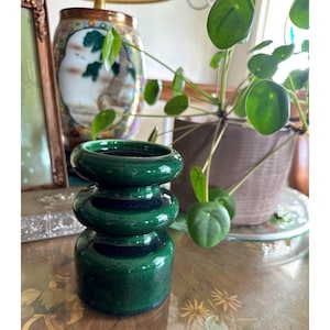 May include: A dark green ceramic vase with a stacked, rounded design. The vase has a glossy finish and a dark interior. The background includes a decorative lamp, a plant, and a vintage table.
