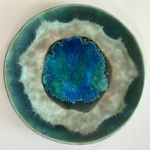 May include: A round ceramic plate featuring a vibrant, crackled cobalt blue center that transitions into a wide, muted beige band, finished with a dark teal outer rim. The glaze displays a textured, crystalline appearance throughout the surface.