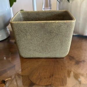 May include: A square, speckled, olive-green ceramic planter. The planter has a slightly tapered shape and a smooth, glossy finish. The interior is empty. The planter is sitting on a wooden surface.