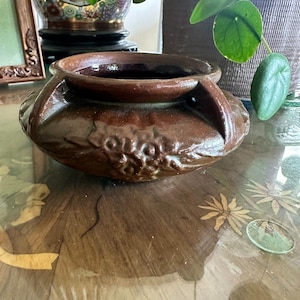 May include: A brown ceramic pot with a wide, rounded body and two handles. The pot has a textured surface with a decorative pattern around the base. The interior is a darker shade of brown.