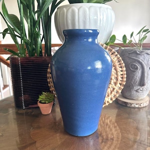 May include: A medium-sized, solid blue ceramic vase with a smooth, matte finish. The vase features a tapered body that narrows toward the base and a slightly flared rim at the top. The surface is uniform in color, showing no patterns or textures.